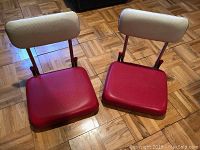 Two portable stadium seats with red padded seats and beige padded backrests on a parquet floor.
