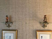 Pair of the figurines mounted on two brass-bracketed glass shelves fixed on a wall above framed artwork.