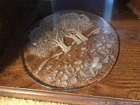 Top view of round clear glass platter showing detailed embossed landscape scene with trees and wildflowers.