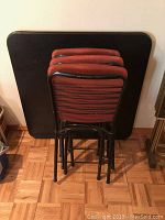 Photo showing four red padded folding chairs stacked in front of the black folding card table, square top.