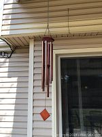 Full view of the brown metal wind chime hanging outside against siding and a window.