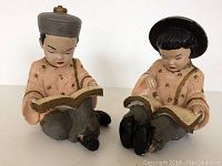 Two vintage plaster figures seated, each reading a book, one with grey cap, the other with black hat, shown from the front.