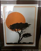 Full view of the framed print showing tree silhouette against an orange sun with reflective glass surface