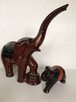 Large brown pottery elephant with a raised trunk, green glaze on back, front foot on stump, glossy finish, shown next to smaller black elephant.
