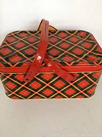 Front view of a vintage tin picnic basket with red and green plaid pattern and red metal handles crossed on top.