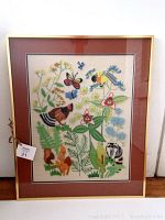 Front view of the framed needlework with nature scenes including birds, butterflies, raccoon and squirrel, flowers, and mushrooms within a gold frame and double matting.