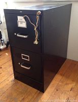 Black two-drawer metal filing cabinet with lock and key hanging on string, placed on wooden floor.