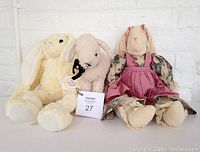 Photo showing three handcrafted plush animals: a large seated bunny doll in a pink dress, a smaller bunny plush with a black ribbon and heritage tag, and a cream-colored lamb plush.