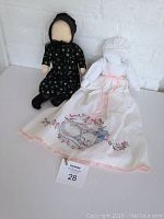 Photo of two handmade Amish Mennonite dolls standing upright on white surface against white brick wall. One doll in black floral dress and bonnet, the other in long white dress with embroidered kitten and bonnet.