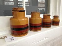 Four vintage ceramic canisters in a honey mustard color with red, black, and brown stripes, each topped with a cork stopper, arranged by size from largest (7") to smallest (4") on a white window ledge.