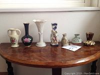 Seven miniature vases arranged on a wooden half-moon table in front of a window with light wall background. Vases vary in shape, size, glazing, and decoration. Included an empty soda can for scale (not in lot).