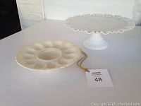 Photo showing both cream-colored devilled egg plate and white cake stand on table with auction tag number 48