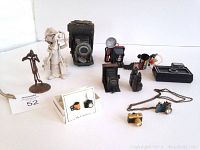 Photo showing entire lot of vintage photography-related collectibles including pottery figurine, miniature cameras, necklace, earrings, pencil sharpeners, water gun, and ornaments.