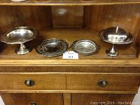 Four serving pieces displayed on a wooden surface: two footed silver-tone dishes (one with lid), one circular silver-tone dish, and one small glass dish.