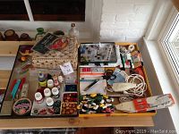 Overall view of wicker sewing basket filled with sewing and craft supplies including threads, needles, buttons, fabric paints, and two electric scissors.