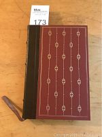 Front cover of the book showing reddish-brown cover panel with vertical gold tooling and brown leather spine with attached fabric bookmark.