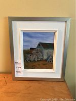 Framed oil painting showing a coastal scene with lobster traps and a building, hung on a beige wall with a MaxSold lot sticker attached to the frame.