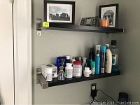 Two photos showing two IKEA black wooden wall-mounted shelves with stainless steel brackets installed on a wall, holding various personal care items and decorative photos.