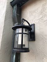 Front view of outdoor wall lantern light fixture mounted against stucco wall showing metal frame, glass cylinder and inner diffuser.