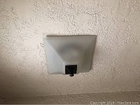 Square ceiling light fixture mounted on textured white ceiling; frosted glass and dark metal finial visible from below.