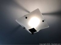 Photo of ceiling light fixture illuminated from within, showing frosted glass square shade with gentle curves and black metal hardware on edges.