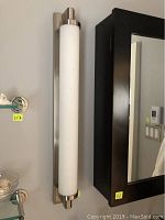 Vertical wall-mounted light fixture showing brushed nickel ends and frosted cylindrical glass shade mounted beside a black framed mirror and glass shelves.