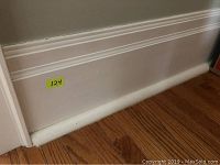 Photo of painted white baseboard molding with a decorative trim near the top, mounted on a wall above a hardwood floor. Yellow label '124' visible.