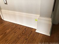Photo of white painted 11-inch high baseboard trim installed along hardwood floor and wall, showing stepped profile and base corner piece.