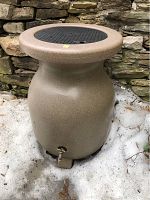 Rain barrel standing on stone patio against stone wall.