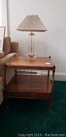 Full view of wooden end table showing the flat top surface with a lamp on it, lower shelf, and one drawer front. Table is next to a couch on a green carpet.