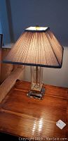 Lamp with clear glass rectangular base and beige pleated tapered shade, placed on wooden table beside upholstered chair.