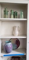 Full view of shelf with four green candle stick vases on top shelf, white pitcher in middle shelf, and purple vase with plate on bottom shelf.
