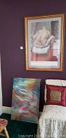 Framed print of a nude figure on a chair, mounted on a purple wall with golden frame, with abstract canvas painting leaning against the wall below it.