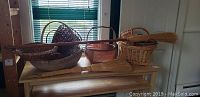 Photo showing eight woven baskets and two natural straw brooms arranged on a wooden table in front of a window.