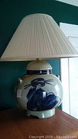 Front view of ceramic table lamp showing blue and white Asian inspired figure on bulbous base with beige pleated shade.