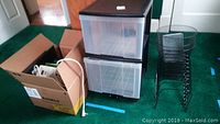 Wide shot showing two-drawer plastic storage next to a cardboard box with power strip, binders and folders, and three black wire mesh wastebaskets