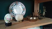 Shelf display with bird-shaped brass candy dish, two brass deer figurines, painted egg on stand, white candle on brass holder, and floral plate behind