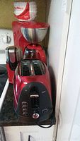 Photo of the red Oster 2-slice toaster in front, KitchenAid coffee grinder on the left, and Hamilton Beach coffee maker behind them, all placed on a kitchen counter.