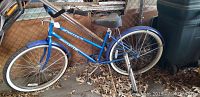 Blue women's Schwinn cruiser bicycle leaning against a chain link fence with leaves on the ground, showing full side profile of bike.