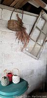 Overview of six pane wooden window with weathered paint leaning against wall, hanging wicker basket with dried flowers, and three ceramic candle holders on nearby surface