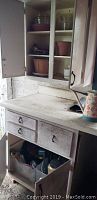 White wooden cabinet with gardening supplies including tools, fertilizers, plastic planters and watering can on counter.