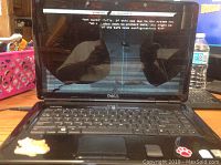 Front view of Dell laptop open, showing cracked and heavily distorted screen with visible error message, keyboard and trackpad visible with some wear.