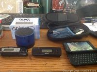 Lot on wooden surface showing two older cell phones, a blue portable Bluetooth speaker, black zippered pouch with cables, and possibly a USB device.