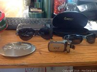 Lot arranged on a desk showing two pairs of men's sunglasses, two belt buckles, and one sunglasses case.