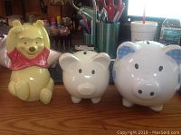 Three piggy banks displayed on table including Winnie the Pooh and two white with blue accent pigs.
