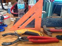Photo of red-handled pliers, yellow utility knife, and orange plastic measuring square on wooden table.