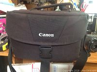 Black Canon-branded camera bag closed with front buckle, showing overall size and material.
