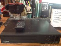 Black Sanyo DVD player with remote on top, showing front loading disc tray and control buttons, placed on a surface with various background items.