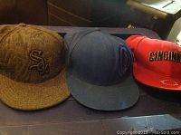 Three baseball hats placed side by side on a flat surface; brown Chicago White Sox hat on the left, blue hat in the middle, red Cincinnati Reds hat on the right.