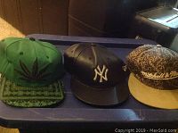 Three men's caps on a plastic storage bin: green cannabis leaf cap, black leather Yankees cap, brown Mitchell & Ness cap